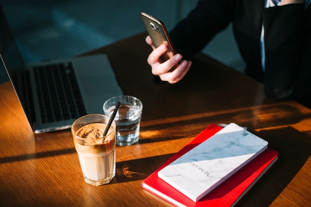 La mano della donna di affari che per mezzo del cellulare con il computer portatile; frappè al cioccolato e libri sulla scrivania in legno