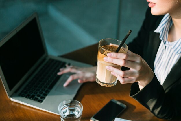 La mano della donna di affari facendo uso del computer portatile e tenendo vetro del frappè del cioccolato