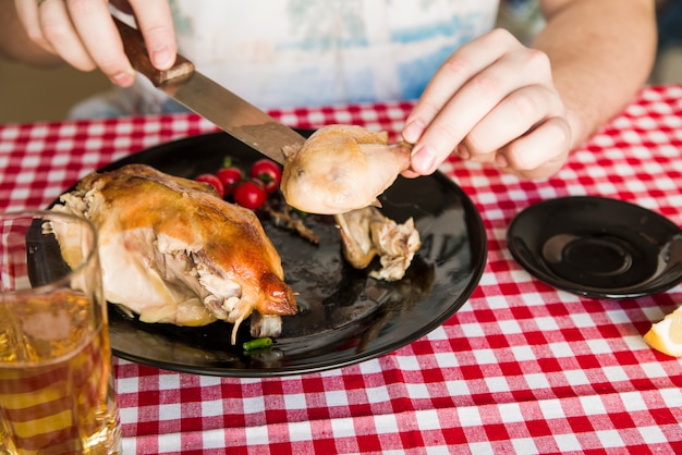 La mano della persona che mangia pollo grigliato facendo uso del coltello mentre cenando con il vetro di birra sul tavolo