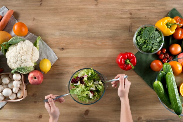 La mano della persona che prepara insalata sana fresca vicino alla varietà di verdure e di frutti sul contatore di cucina di legno