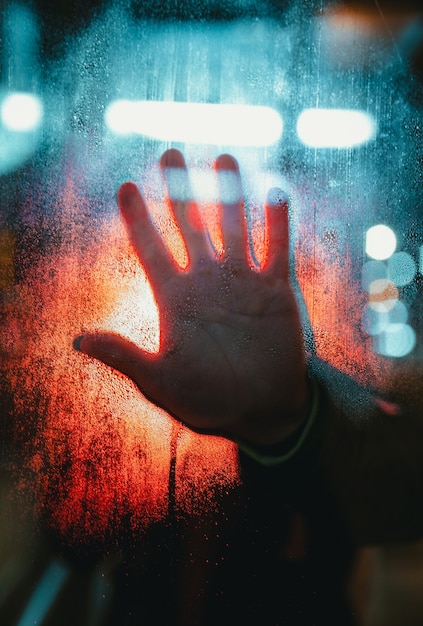 La mano della persona che tocca un vetro coperto di gocce di pioggia con luci bokeh