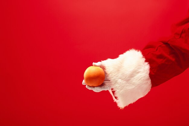 La mano di Babbo Natale che tiene un frutto arancione su sfondo rosso. La stagione, l'inverno, le vacanze, la celebrazione, il concetto di regalo