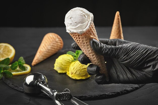 La mano in un guanto scuro tiene in mano un cono di cialda con un delizioso gelato cremoso sullo sfondo di un sorbetto al limone decorato con menta e coni di cialda che vengono serviti su un'ardesia di pietra su un bac nero