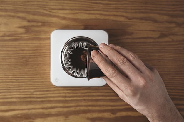 La mano maschile versa caffè macinato tostato dalla tazza in metallo inossidabile alla caffettiera a goccia cromata per preparare il caffè filtrato nella caffetteria