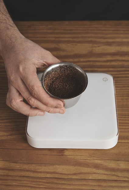 La mano maschio tiene la tazza d'argento inossidabile con caffè macinato tostato sopra i semplici pesi bianchi su un tavolo di legno spesso. vista dall'alto