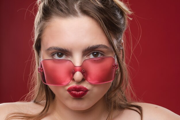 La modella caucasica con lunghi capelli biondi e grandi occhiali da sole rossi posa per la macchina fotografica e si rallegra