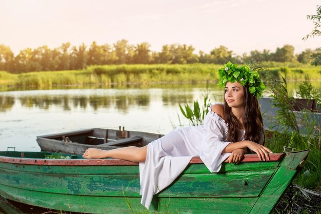 La ninfa dai lunghi capelli scuri in un abito bianco vintage seduta su una barca in mezzo al fiume. Tra i capelli una corona di foglie verdi con fiori bianchi. Servizio fotografico di fantasia.