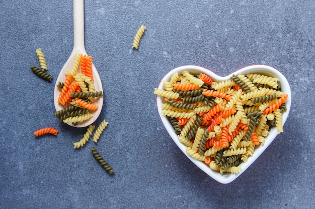 La pasta variopinta dei maccheroni in un cuore ha modellato la ciotola e il cucchiaio su una superficie grigia