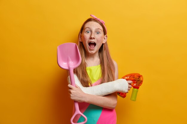 La pazza e giocosa ragazza rossa tiene la bocca aperta, gioca con la pistola ad acqua durante le calde giornate estive, si diverte in riva al mare, tiene in mano una pala giocattolo di sabbia di plastica, indossa il costume da bagno, si diverte con i divertenti giochi estivi con gli amici