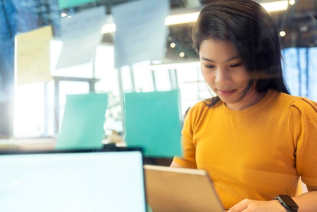 La persona creativa femminile asiatica freelance attraente indossa un panno giallo casual che lavora con il computer portatile nell'area di condivisione dello spazio di coworking nuovo stile di vita con l'emozione pensierosa e fresca sfocatura dello sfondo dell'ufficio