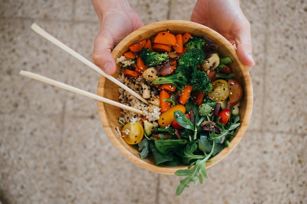 La persona vegana regge una ciotola di buddha in legno di cocco piena di verdure, verdure, cereali e bacchette sani.