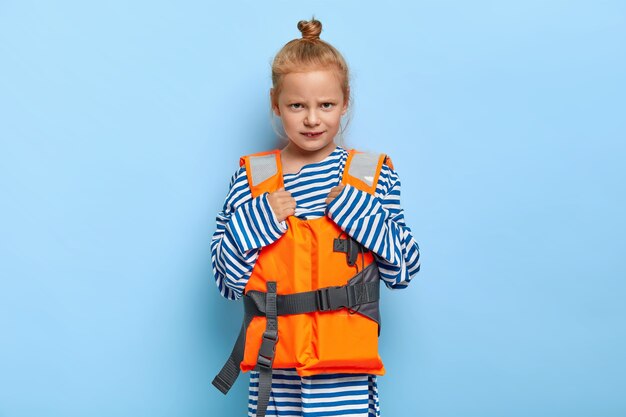 La piccola bambina arrabbiata con il panino dei capelli di zenzero riposa durante le vacanze estive indossa un maglione a righe oversize ei genitori dispiaciuti più vivaci non le permettono di nuotare da sola con l'aiuto del nuoto. Ragazza in giubbotto di salvataggio