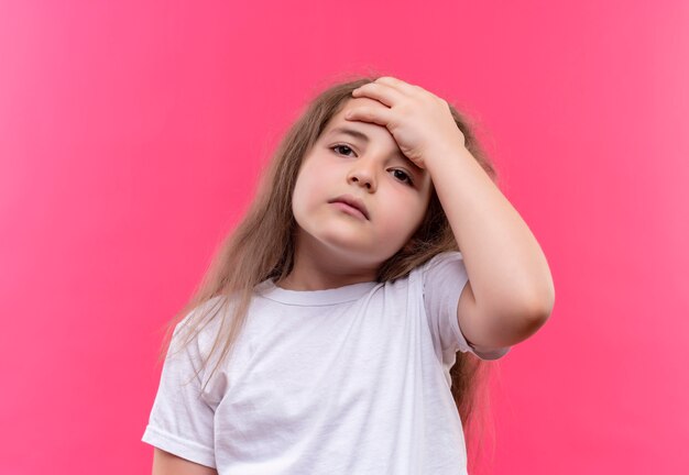 La piccola ragazza ammalata della scuola che porta la maglietta bianca ha messo la sua mano sulla fronte su fondo rosa isolato