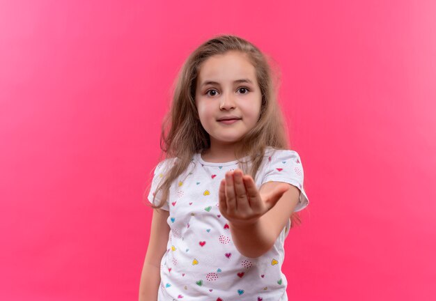 La piccola ragazza sorridente della scuola che porta la maglietta bianca tese la mano su fondo rosa isolato