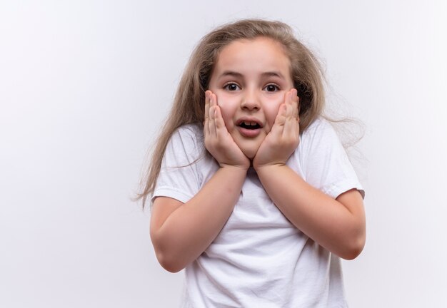 La piccola ragazza spaventata della scuola che porta la maglietta bianca ha messo le sue mani sulle guance su fondo bianco isolato