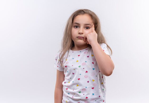 La piccola ragazza triste della scuola che porta la maglietta bianca ha messo il suo dito sull'occhio su fondo bianco isolato