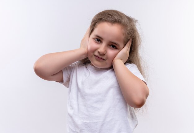 La piccola ragazza triste della scuola che porta la maglietta bianca ha messo le sue mani sulle orecchie su fondo bianco isolato