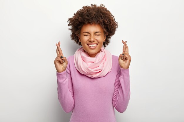 La promettente donna dai capelli ricci intensi solleva le dita incrociate, concentrata sul sogno, anticipa il risultato positivo, sorride ampiamente, indossa abiti viola, sta contro il muro bianco, supplica per buona fortuna