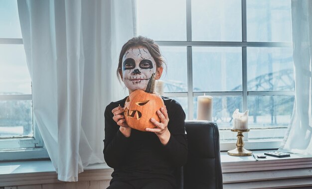 La ragazza abbastanza spaventosa si sta preparando per Halloween in un appartamento vicino alla finestra, tiene in mano la zucca.