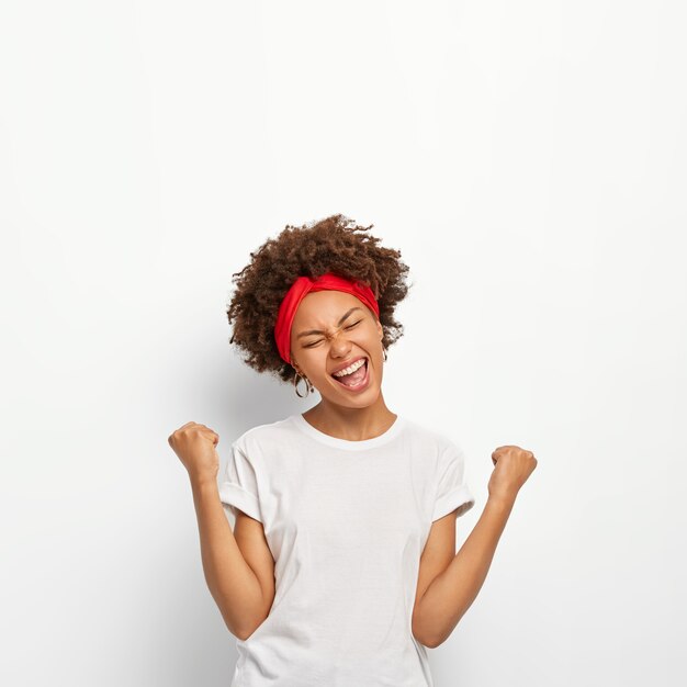 La ragazza afro felice e felice stringe i pugni, si sente trionfante, si rallegra della vittoria, tiene gli occhi chiusi, sorride ampiamente, indossa una maglietta bianca, sta al coperto