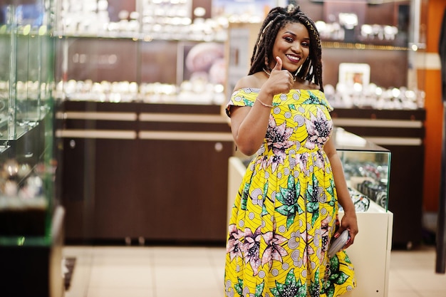 La ragazza afroamericana carina di piccola altezza con i dreadlocks indossa un vestito giallo colorato sul negozio di orologi al centro commerciale