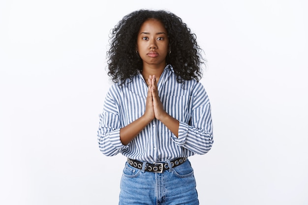 La ragazza afroamericana dall'aspetto serio ha bisogno di aiuto pregando tenendosi per mano pregare supplicando, i palmi premuti insieme per favore fai il favore, implorando in piedi cupo intenso muro bianco preoccupato