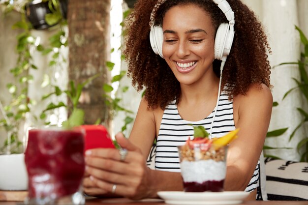 La ragazza afroamericana positiva dei pantaloni a vita bassa cerca nuove canzoni di musica, felice di ricevere un messaggio sul cellulare. L'amante della musica femminile ascolta la composizione dalla playlist, digita gli sms di testo nei social network