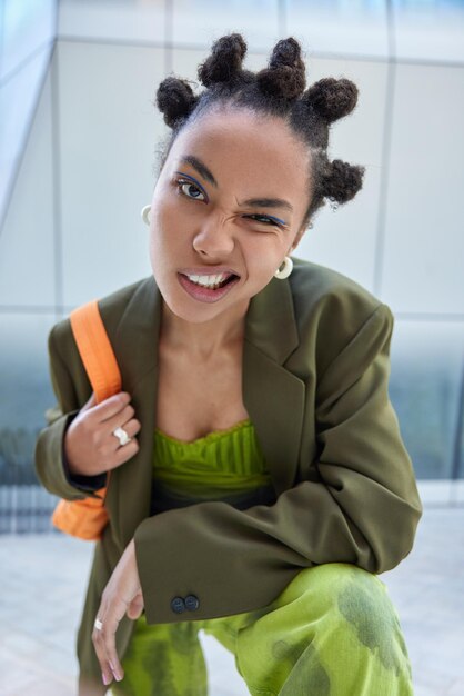 La ragazza alla moda hipster strizza l'occhio agli occhi fa un'espressione sfacciata vestita in costume alla moda verde porta la borsa ha un trucco vivido di acconciatura da ciambella posa all'aperto. Stile di vita urbano