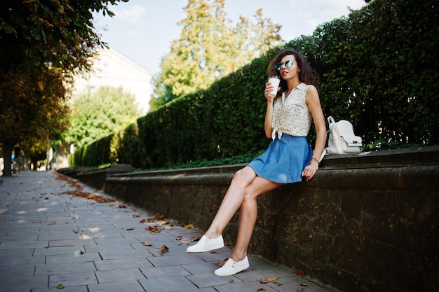 La ragazza alla moda riccia indossa la camicetta del pannello esterno dei jeans blu e gli occhiali posati sulla strada della città con una tazza di caffè