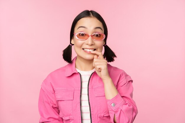 La ragazza asiatica alla moda in occhiali da sole sembra premurosa o incuriosita vuole provare smth out stand su sfondo rosa riflettendo