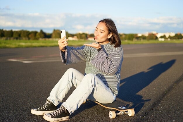 La ragazza asiatica felice si siede sullo skateboard prende selfie con il longboard fa facce carine giornata di sole all'aperto