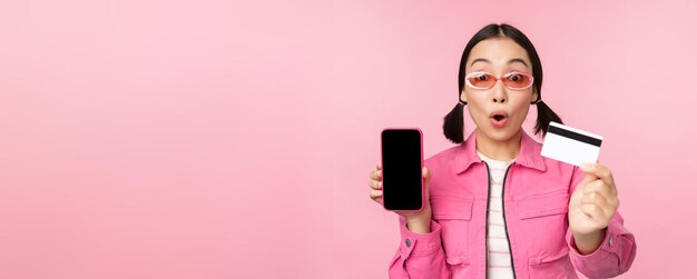 La ragazza asiatica mostra lo schermo del telefono cellulare e la carta di credito reagisce sorpresa dalla fotocamera che boccheggia impressionato in piedi sopra il concetto di shopping online di sfondo rosa