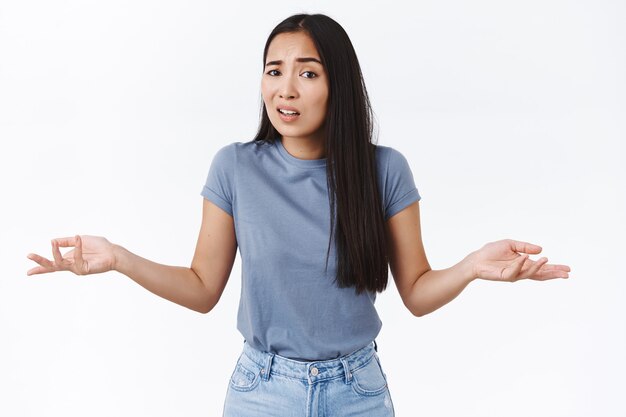 La ragazza asiatica non ha capito cosa sta succedendo, in piedi perplessa, cosa vuoi da me in posa, alzando le spalle con le mani aperte lateralmente per lo sgomento, non ti aiuterà, indifferente al tuo problema