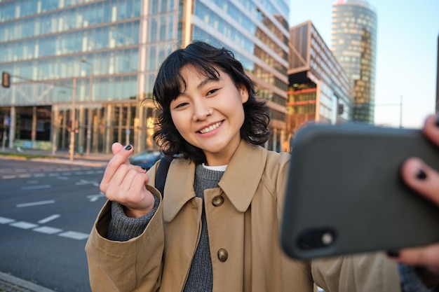 La ragazza asiatica positiva prende il selfie sullo smartphone mostra il cuore con le dita e fa una foto sulla strada
