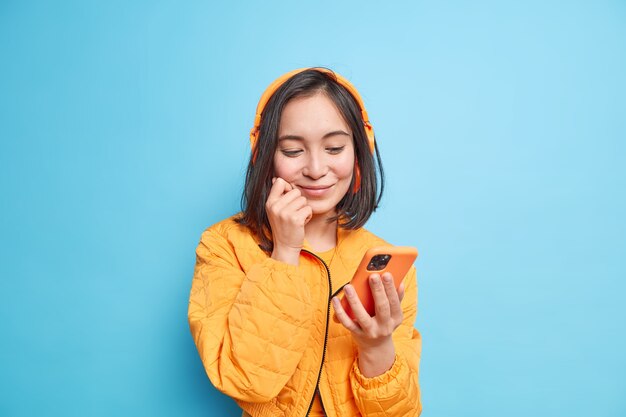 La ragazza asiatica soddisfatta con i capelli scuri concentrata sul display dello smartphone sceglie la traccia audio dalla playlist indossa cuffie stereo wireless sulle orecchie vestite con una giacca arancione isolata sul muro blu