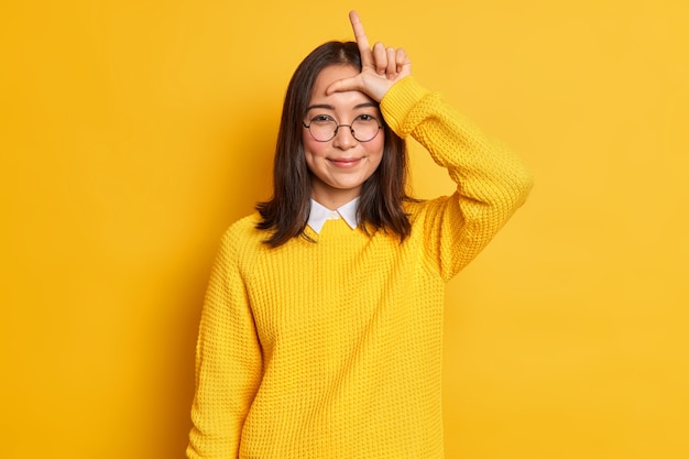 la ragazza asiatica sorridente fa il gesto del perdente sulla fronte guarda con soddisfazione dice che l'amico hai perso la forma La lettera L con le dita prende in giro la squadra indossa occhiali rotondi e maglione