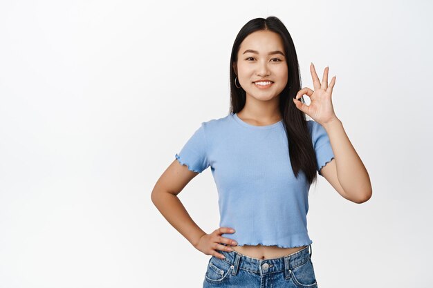 La ragazza asiatica sorridente mostra il gesto giusto conferma qualcosa di dire di sì facendo il segno zero di Ok in piedi su sfondo bianco