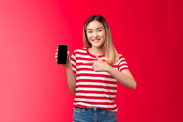 La ragazza bionda asiatica carina allegra molto soddisfatta che sorride ampiamente mostra il display dello smartphone che punta il telefono...