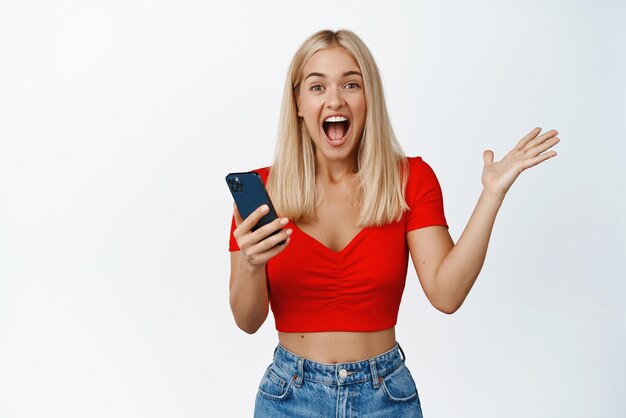 La ragazza bionda eccitata reagisce alla notifica del telefono cellulare tenendo lo smartphone e gridando di gioia in piedi su sfondo bianco