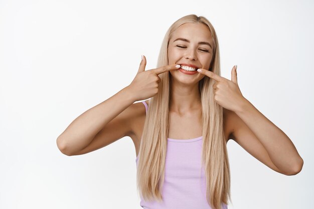 La ragazza bionda felice sorride punta il dito contro i suoi denti bianchi e l'espressione del viso soddisfatta si erge su sfondo bianco