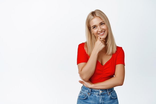 La ragazza bionda glamour sorride sembra civettuola alla telecamera con top corto rosso e jeans su sfondo bianco