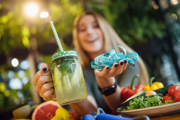 La ragazza bionda si siede sul tavolo e tiene in mano un frullato fresco e misura, concetto di tempo di dieta.