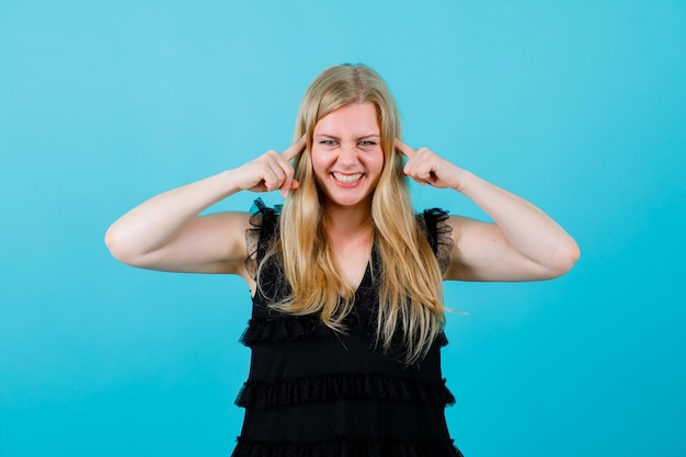 La ragazza bionda sorridente si copre le orecchie con gli indici su sfondo blu