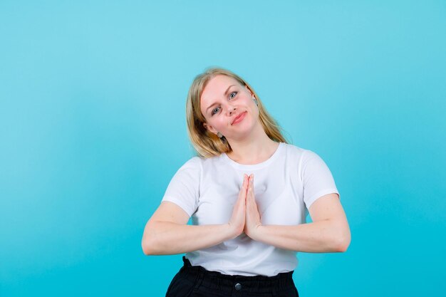 La ragazza bionda sta chiedendo l'elemosina tenendo le mani insieme sul petto su sfondo blu