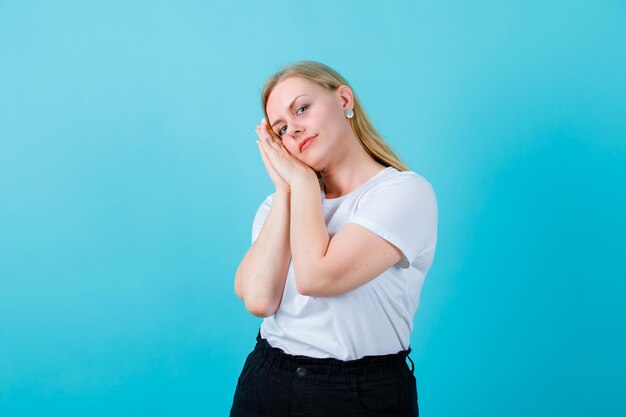 La ragazza bionda sta facendo il gesto del sonno tenendo la testa sulle mani su sfondo blu