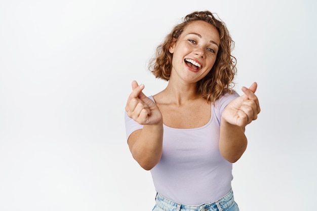 La ragazza bionda sveglia con i capelli corti mostra i cuori del dito, ridendo e sorridendo, fondo bianco