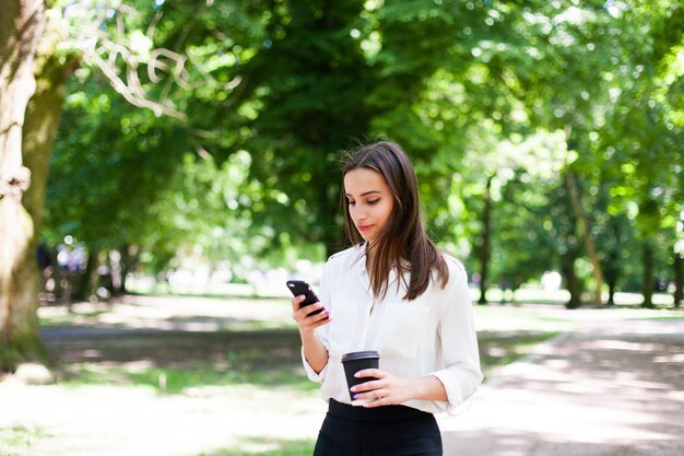 La ragazza cammina con il telefono in mano e una tazza di caffè nel parco