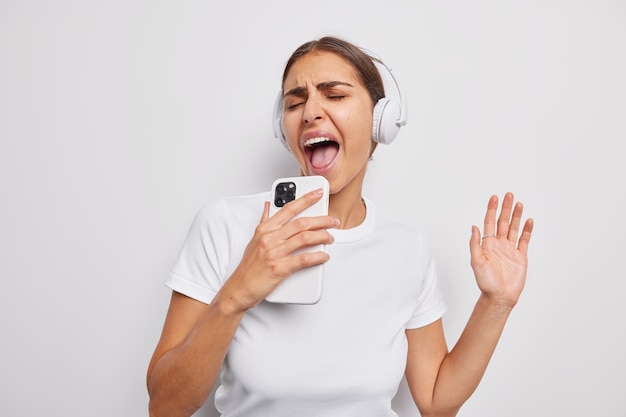 la ragazza canta la canzone ascolta la musica nelle cuffie tiene il telefono cellulare vestito casualmente ha un'espressione allegra su bianco