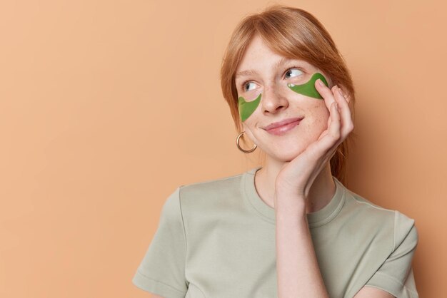 La ragazza carina e sognante pensierosa dello zenzero ha un'espressione sognante distoglie lo sguardo applica patch di bellezza verdi per rimuovere le linee sottili e le borse sotto gli occhi vestita con una maglietta casual isolata su sfondo marrone