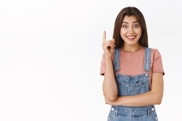 La ragazza carina sollevata finalmente ha avuto un'idea eccellente, condividendo il suo piano o suggerimento, sorridendo felice e sciocca macchina fotografica, alzando un dito nel gesto di eureka, ottenendo una soluzione, in piedi sfondo bianco